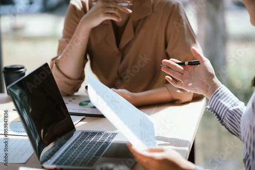 Close-up of two businesswomen working on a laptop, one pointing at the screen while the other types, showing teamwork, guidance, and collaboration in a bright office.