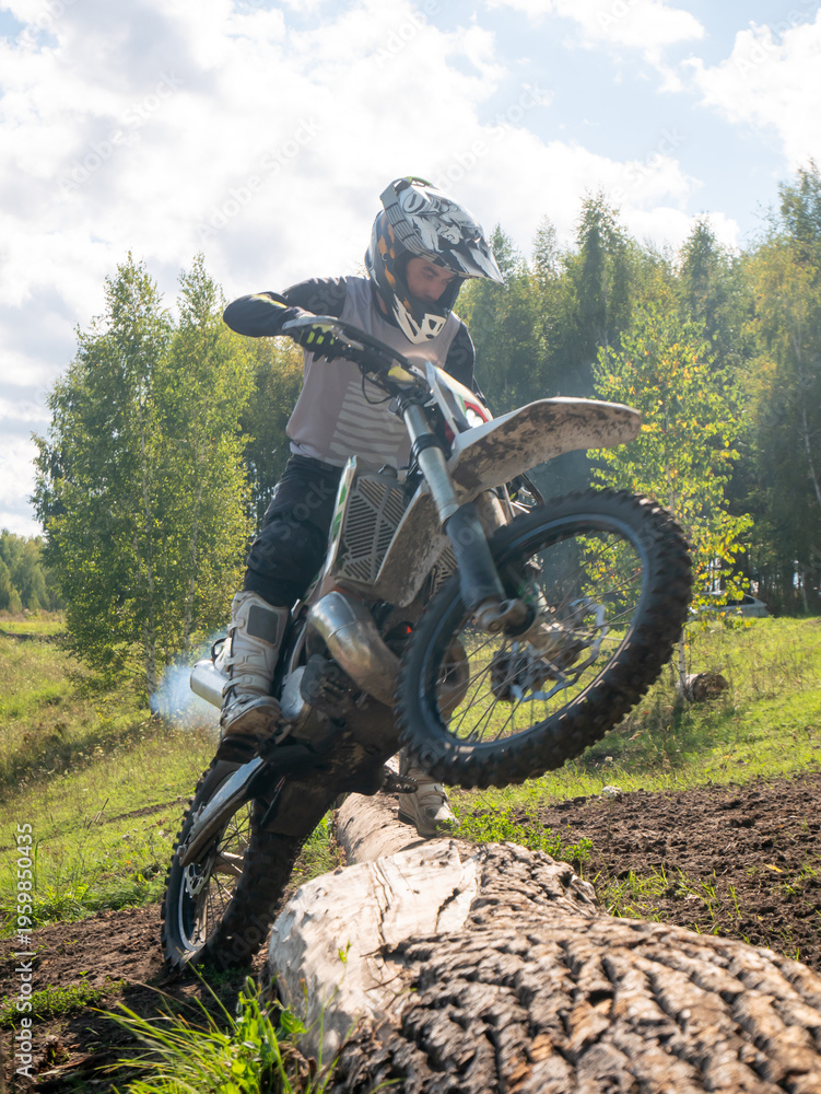 Obraz premium Motocross rider performing wheelie over fallen tree log in forest. Man racing dirt bike on off-road terrain. Extreme sports activity, speed, adrenaline, skill and outdoor adventure ride.