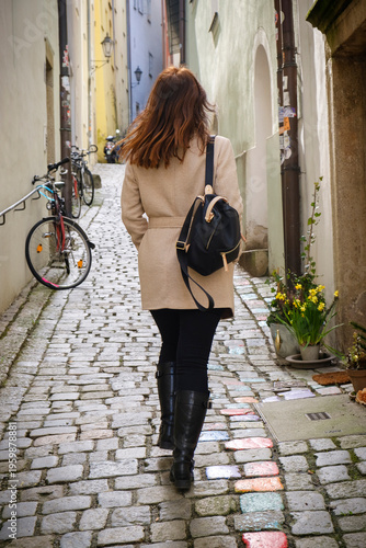 Back view of woman walking along narrow European street with cobblestones. Urban lifestyle and travel concept.