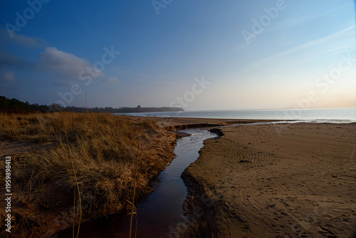 Coastal beach with a winding stream flowing into the sea, dry grass dunes and calm horizon under soft evening light.