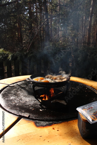 Foto Fried eggs and meat cooking in a cast iron skillet over an open flame on a rustic outdoor grill, featuring rising steam and a blurred forest background