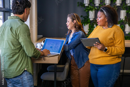 Diverse coworkers meeting at high counter in office in casual clothes, using laptop tablet headset