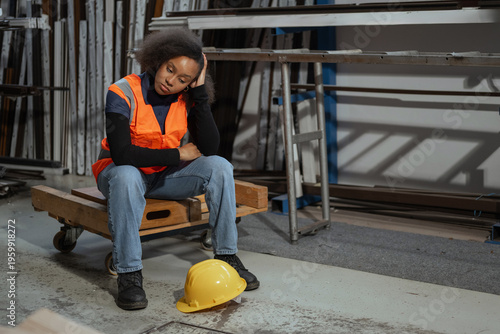 Fatigued African American female warehouse worker taking off a yellow hard hat and sitting down to rest after a hard workday.