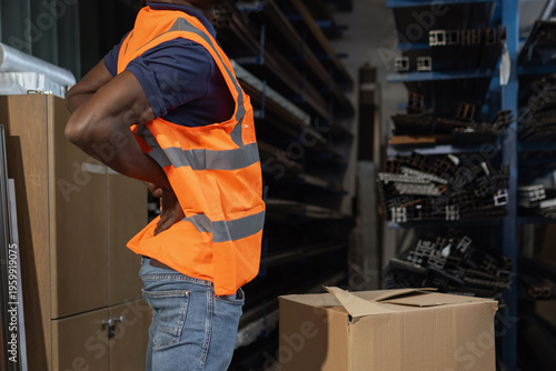 Warehouse worker in a safety vest holding and stretching his painful lower back after carrying and lowering a heavy cardboard box.