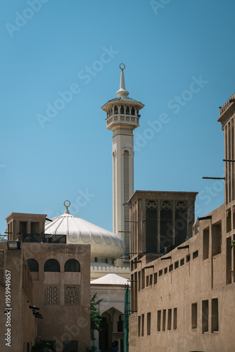 Mosque minaret and white dome rising above traditional wind towers and coral-stone walls in the historic district of Dubai, United Arab Emirates, under a clear blue sky