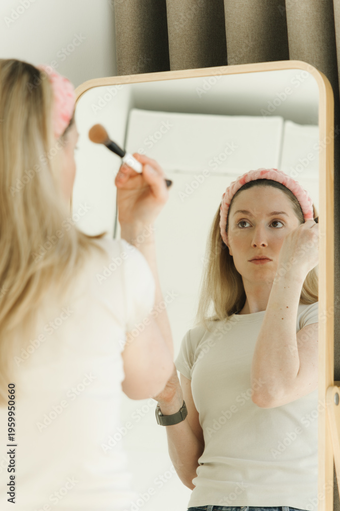 Naklejka premium Young woman applying makeup in front of a mirror at home. Natural beauty routine with soft light, skincare concept, and everyday self care lifestyle.