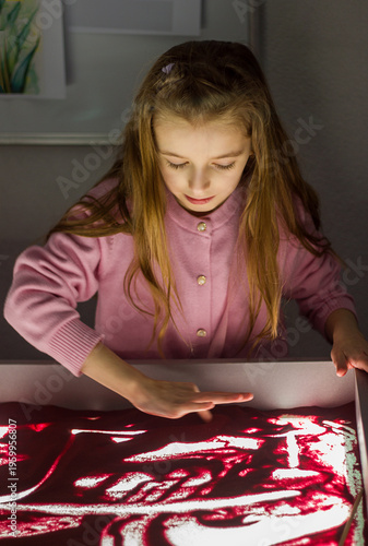 Beautiful little girl draws with sand on a light square, art therapy, sand animation, creative leisure for children, selective focus