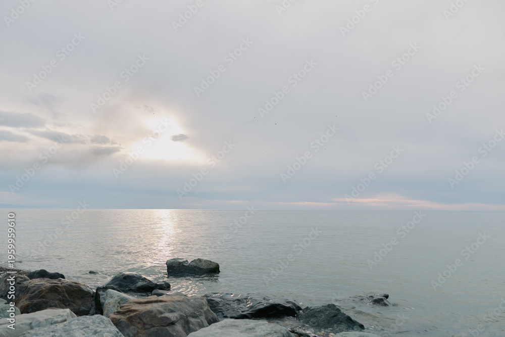 Fototapeta premium Calm sea with gentle waves and rocks in foreground under cloudy sky at sunrise. Peaceful coastal landscape with soft natural light reflecting on water surface.