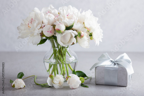 Still life with a bouquet of white peonies in a glass vase, a silver gift box, tied with a satin ribbon on a gray textured surface. Elegant flower arrangement for a birthday or wedding.