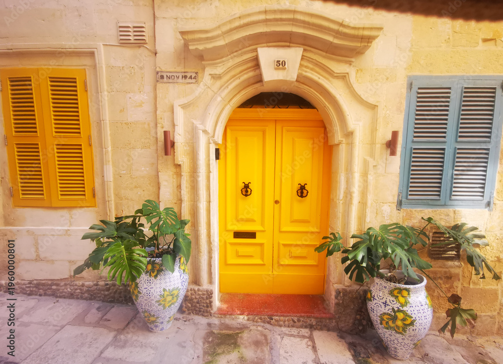 Fototapeta premium Yellow wooden door on a Maltese stone facade framed by decorative plants. The image conveys the concept of architectural detail and colorful Mediterranean style.