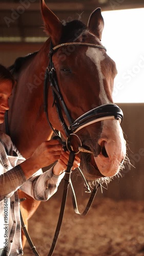Woman shows concern by petting horse in stable, adjusting its bridle. Concept equestrian sport, horse care. 