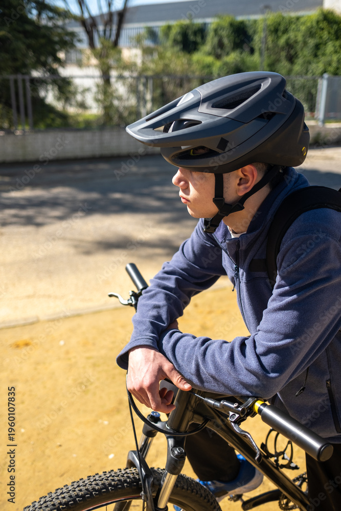 Naklejka premium Young male cyclist wearing a protective helmet and backpack, resting with arms crossed on his mountain bike outdoors