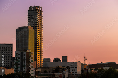 Modern skyscrapers with glass and concrete facades illuminated by golden sunset light against vibrant pink and orange sky in Puebla, Mexico