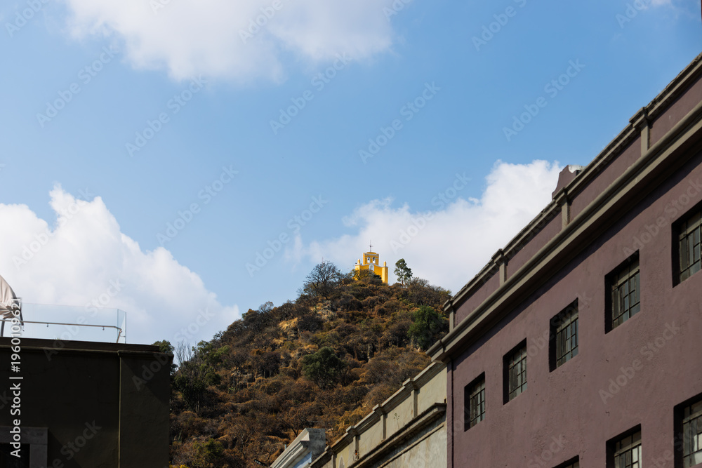 Obraz premium View of yellow Chapel of San Miguel Arcangel atop dry hill overlooking colorful urban buildings under blue sky with clouds in Atlixco, Puebla, Mexico