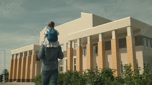 dad carries a little daughter on his shoulders to school, a girl with a school bag on his shoulders, walk around the schoolyard, see off to first grade, preschool education, father day, happy family.
