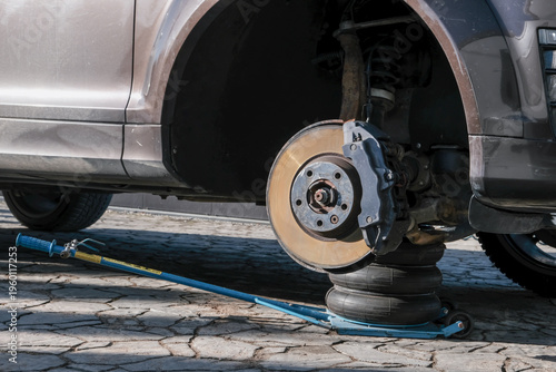 A car brake disc with visible rust and dust, mounted on a vehicle lifted by a blue jack. Brake caliper and mechanical components in focus, high performance car brake system condition checking