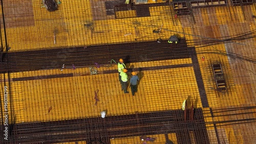 Aerial drone shot looking straight down at workers assembling dense steel rebar grid on formwork deck, highlighting structural reinforcement, teamwork, and building progress.