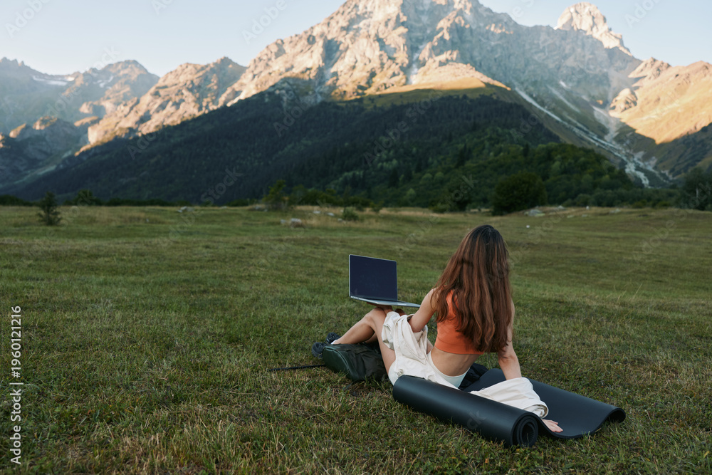 Naklejka premium A person sits on a grassy meadow with a laptop, distant mountains in the background, creating a peaceful outdoor work moment and a sense of calm focus for creative tasks and learning.