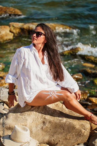 Mature woman in white shirt sitting on rock near ocean with long hair blowing and sun cream spiral on leg