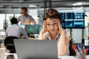 Frustrated female office worker sits at desk, feeling stressed and burned out while working on laptop. Caucasian woman with hands on head, showing signs of exhaustion and tension in busy workplace.