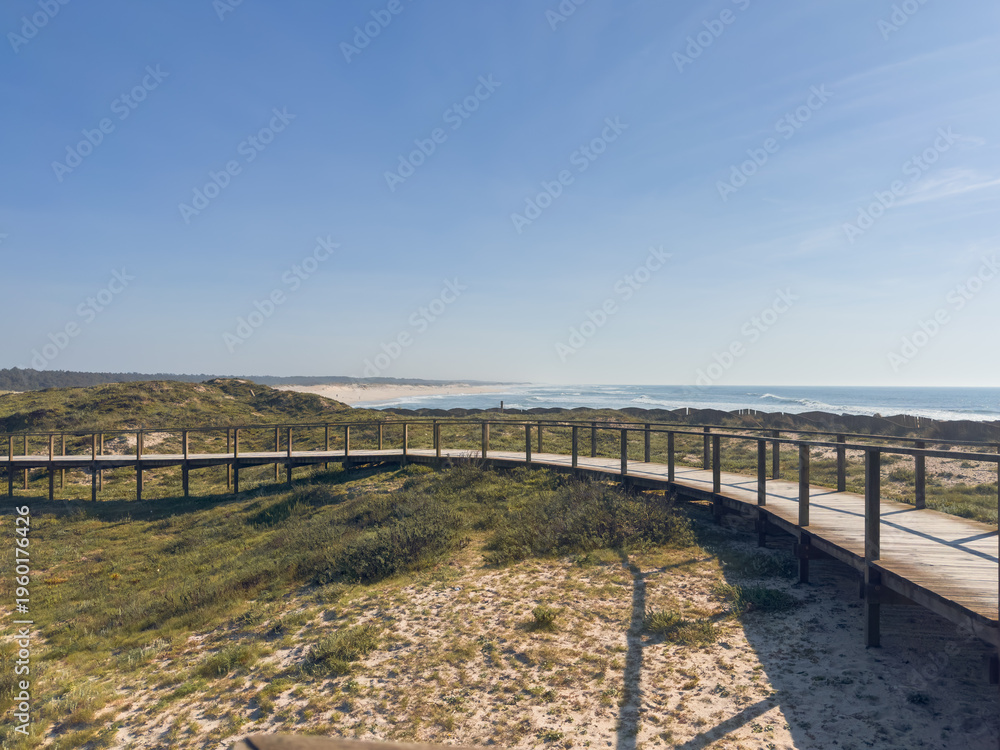 Fototapeta premium Coastal wooden walkway winding through natural sand dunes towards the Atlantic Ocean under a bright blue sky at Furadouro beach in Ovar Portugal