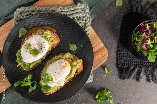 A top down view of two slices of crusty sourdough topped with creamy mashed avocado and perfectly poached eggs with rich golden yolks flowing across a black plate and rustic wooden board