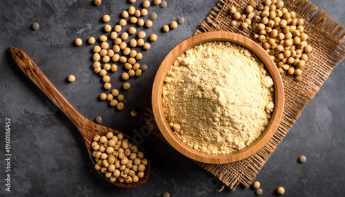 Overhead view of soybeans and soy flour in a wooden bowl with a spoon on a dark surface.