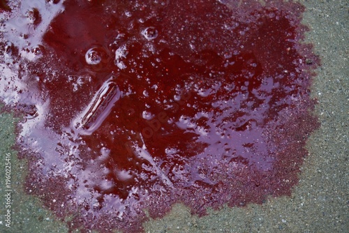 Red liquid spill on rough concrete surface with small bubbles and reflections on top of ground
