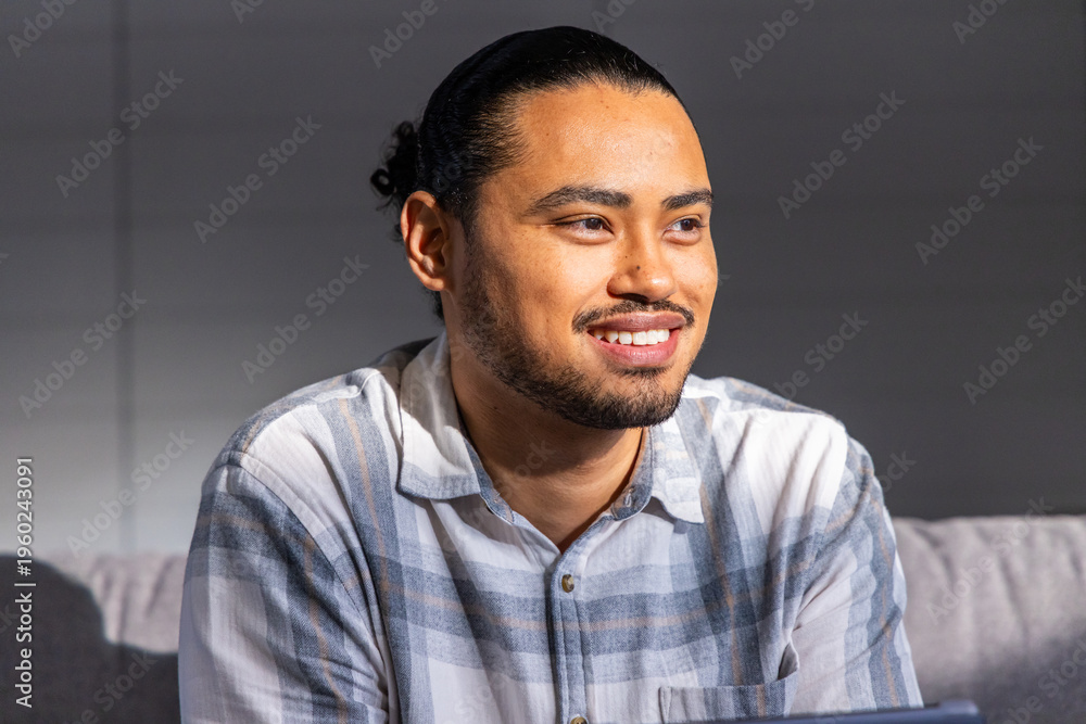 Obraz premium Man sitting on grey upholstered sofa in living room, wearing plaid shirt and using laptop