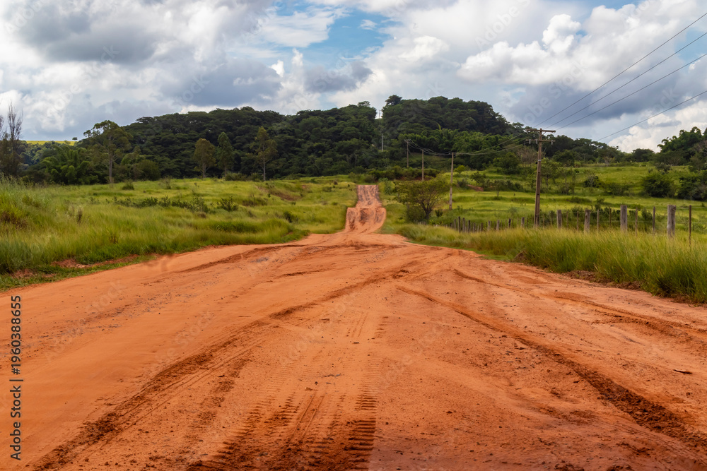 Naklejka premium Red dirt rural road crossing farmland under dramatic storm clouds, with power lines and a lone dry tree, depicting countryside landscape, severe weather and agricultural infrastructure.
