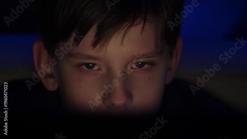 Close-up portrait of a boy staring intently at a computer screen in a dark room. The child's face is illuminated by the soft light from the display against a blue neon background