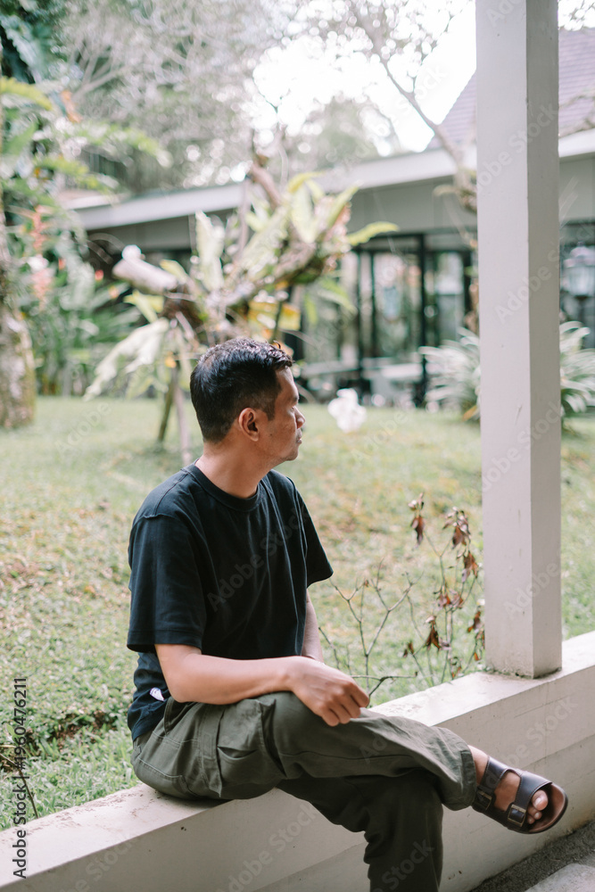 Fototapeta premium Asian Man Sitting and Looking Away Thoughtfully in a Peaceful Garden