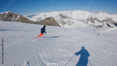 A skier in bright orange pants skis down a snowy mountain slope with snow capped mountains in the background. Slow motion POV	
