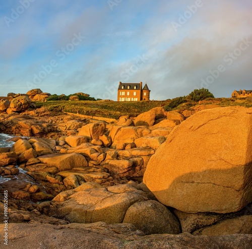 Traditional stone house on a rocky coastline during golden hour. Dramatic seaside landscape with warm sunlight, granite boulders and ocean waves. Coastal scenery in Brittany, France, ideal for travel