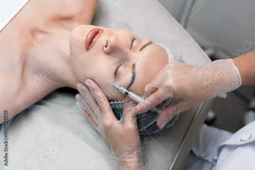 Woman head lying with closed eyes while a dermatologist or aesthetician in gloves performs a facial injection near the eye area in a wellness clinic, close-up.