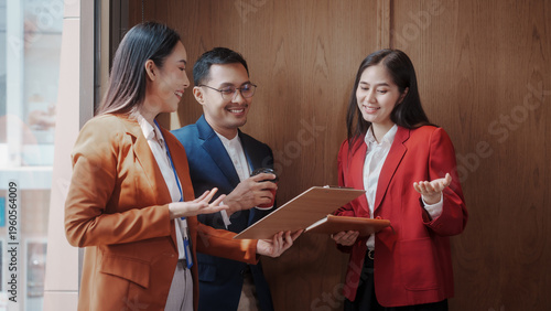 Three business colleagues in suits discuss documents in a modern office hallway, smiling and collaborating during a meeting.