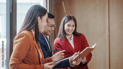 Three business colleagues in suits discuss documents in a modern office hallway, smiling and collaborating during a meeting.