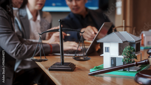 Legal consultation about real estate: scales of justice beside a model house on desk, with business people meeting and using a laptop in the background.