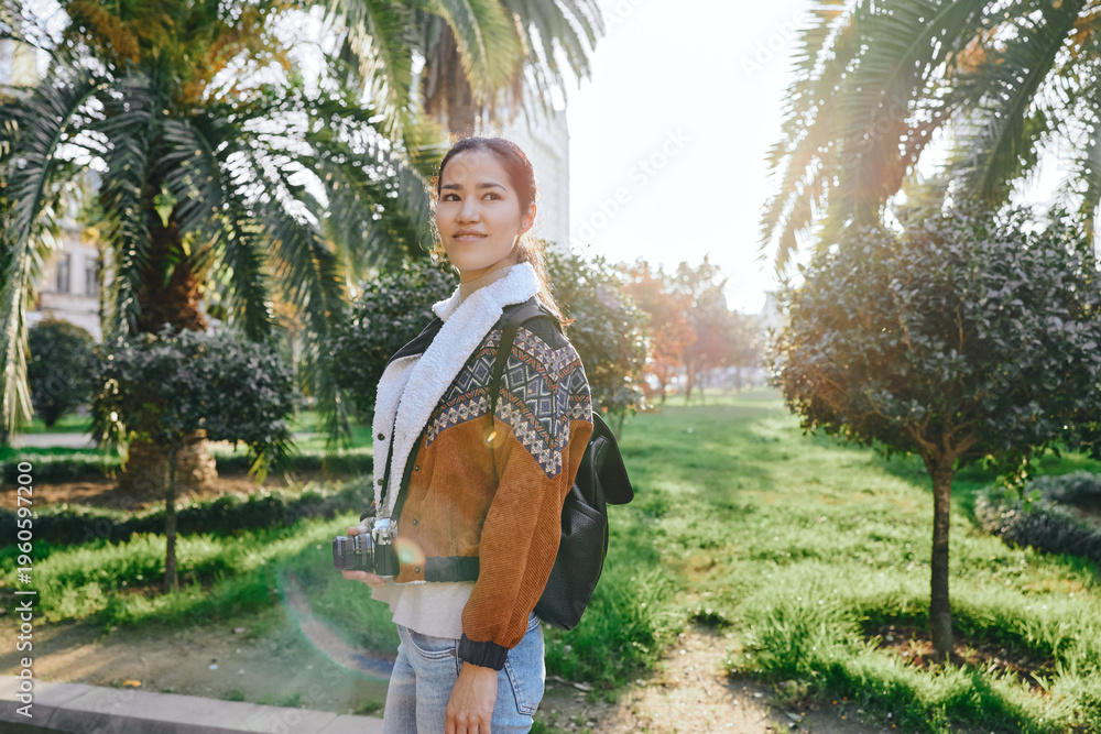 Fototapeta premium Woman with camera in sunny park, outdoor fashion, backpack, casual outfit, capturing moments amid palm trees and green lawn, bright warmth and relaxed mood