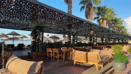 An empty beachfront terrace with wooden furniture and lounge seating under a dappled shade canopy. The view overlooks a calm sea with distant mountains and sunlit palm trees in the background.