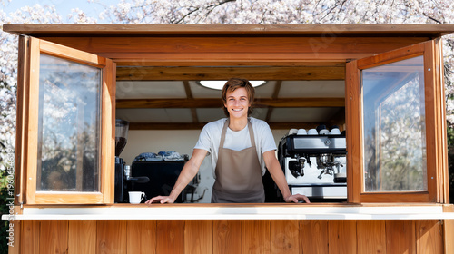 Smiling young male barista in beige apron standing inside wooden outdoor coffee kiosk with espresso machine, cherry blossom trees background, spring cafe season opening.