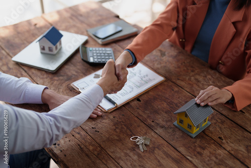 Business partners seal a property deal with a handshake over a wooden desk strewn with house models, keys, calculator and contract, symbolizing real estate transaction success
