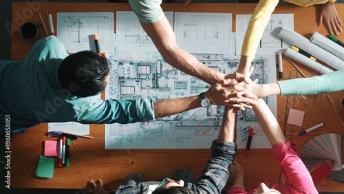 Slika na platnu Top down aerial view of designer putting hand together to celebrate successful project at meeting table with blueprint and project plan place