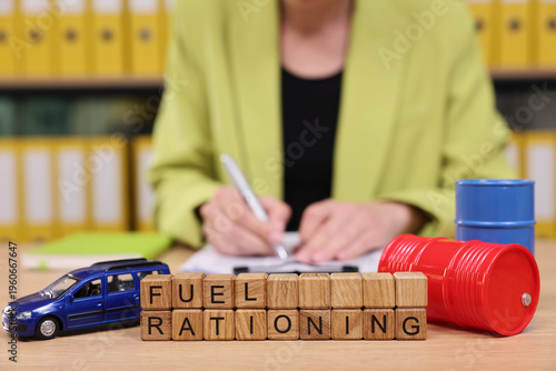 A person in a vibrant blazer sits at a desk, writing notes while surrounded by symbols of fuel rationing. Miniature car and fuel container illustrate the urgent theme of resource management.