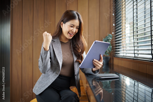 Excited businesswoman celebrating success while using tablet in modern office. Business success and achievement concept.