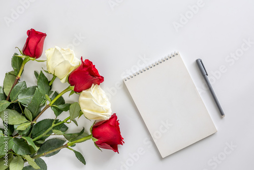 Bouquet of red and white roses, blank white piece of greeting paper with pen on white background. Copy space, flatlay. Concept: Birthday gift, Valentine's Day, Mother's Day and Women's Day