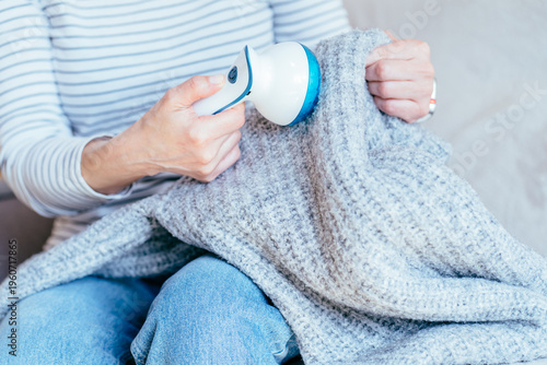 Woman cleaning gray knit blanket with fabric shaver while sitting at home