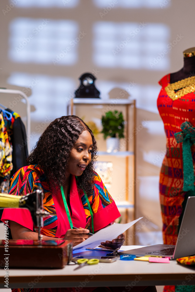 Fototapeta premium African fashion designer managing business, reviewing documents, and using laptop in her studio