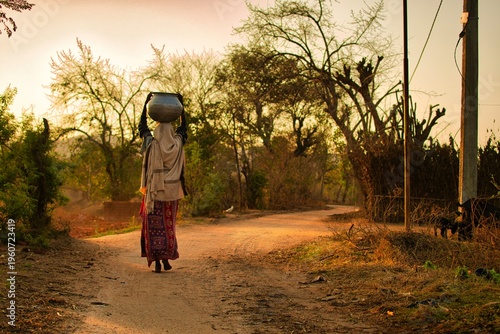 young rural woman walking on the village path in the morning carrying a load on her head