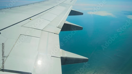 Airplane flying above calm blue sea with wing flaps visible. Jet cruising over ocean waters with wing structure in frame. Passenger aircraft gliding across marine horizon showing wing panels